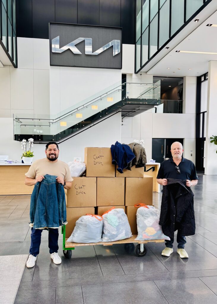 Two men stand on either side of boxes filled with coat donations, each holding a donated coat