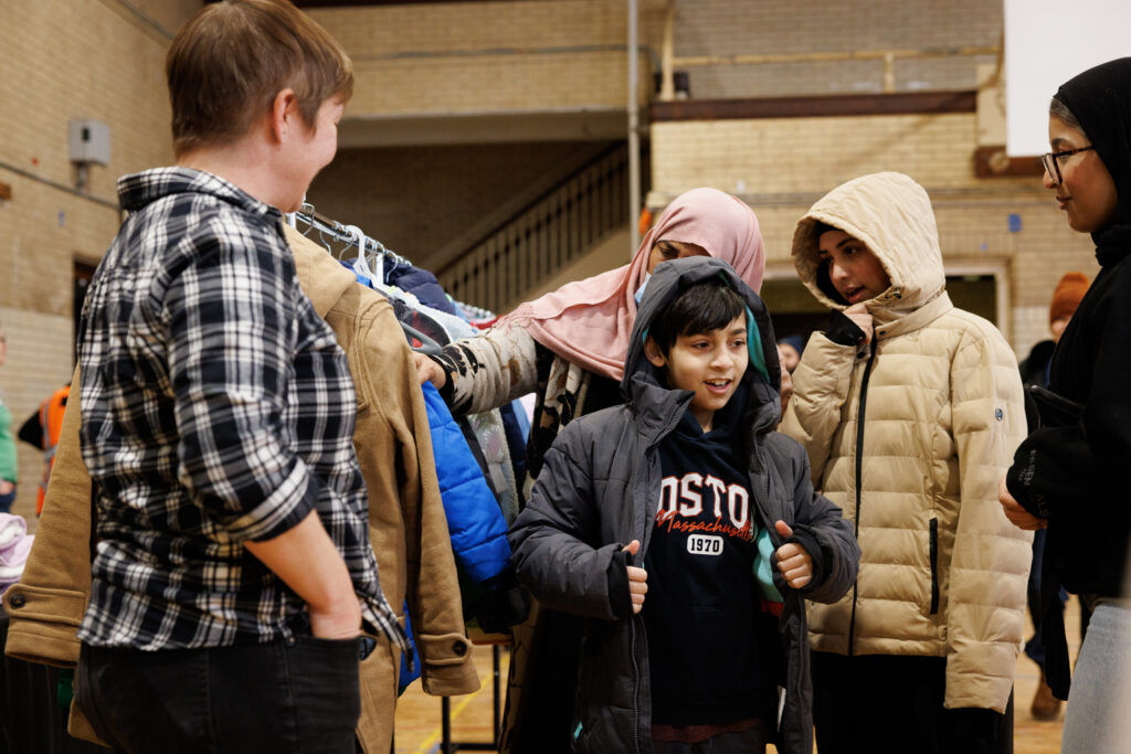 A group of people sharing coats in a community setting. One warm coat impact.