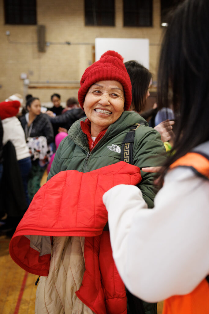 An older woman wearing a red hat happily accepts a new red coat at a coat drive.