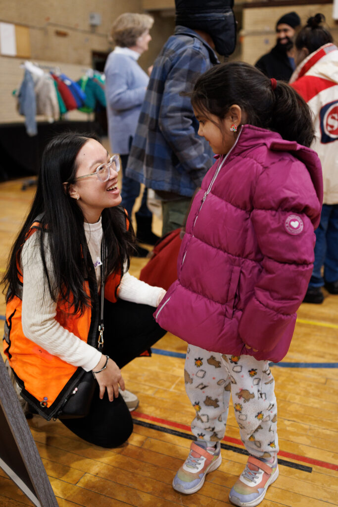 A woman wearing an orange vest puts a purple coat on a young girl.