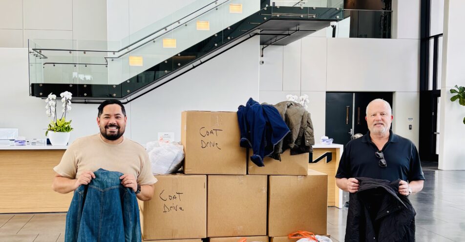 Two men stand on either side of boxes filled with coat donations, each holding a donated coat
