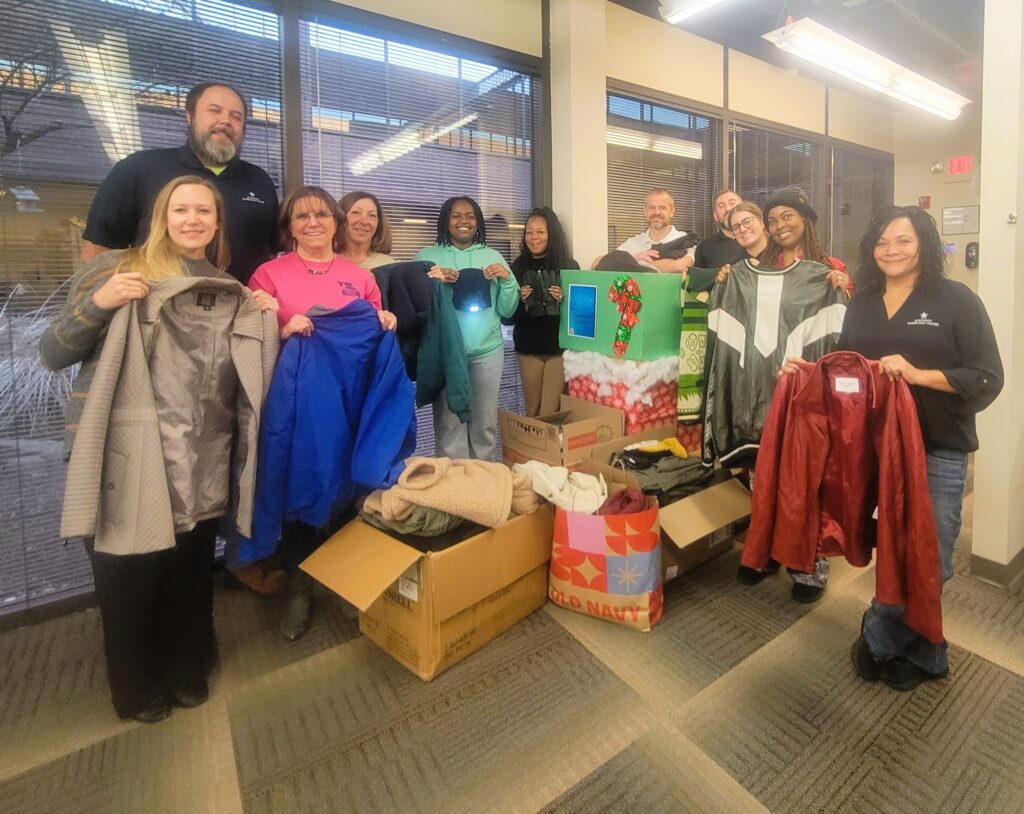 People smiling, holding coats, with boxes of clothing donations. Likely related to one warm coat impact.